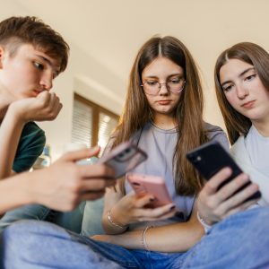 One teenage boy and two teenage girls have fun together using mobile phones at home. Beautiful young people are friends and high school students.