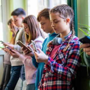 Teenage students are standing in the school hallway, all looking at their phones.